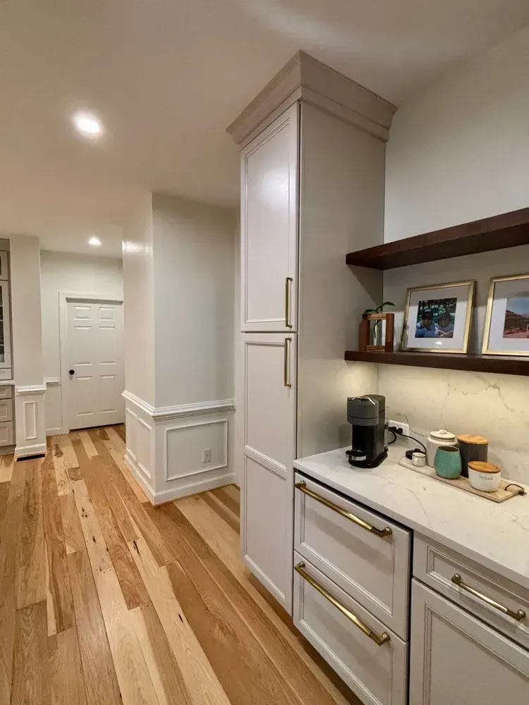 Bright kitchen hallway with white cabinets, wood floor, and countertop items under framed photos.