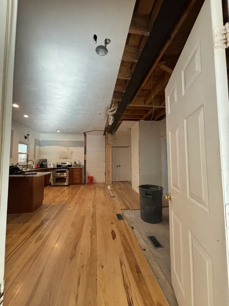 Narrow kitchen and living area with exposed ceiling beams, white walls, wood floor, and open doorway.