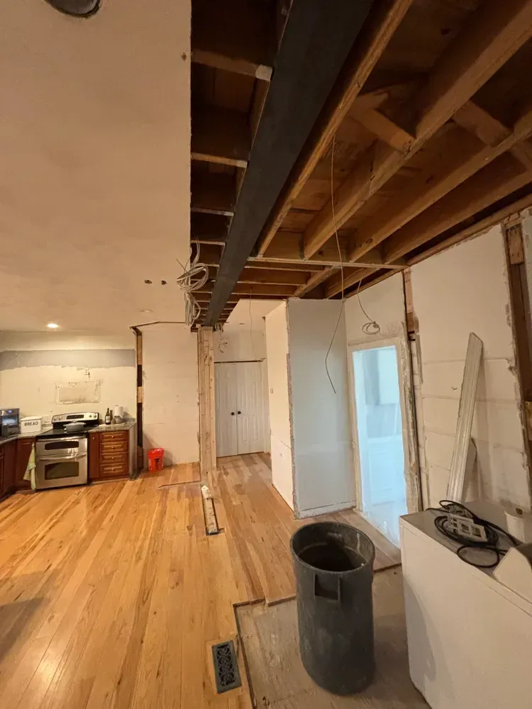 Partially renovated room with exposed ceiling beams, wood floor, white cabinets, and a trash can