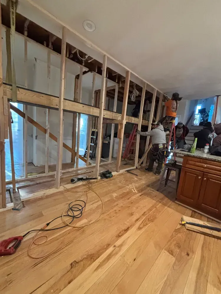 Kitchen under renovation with exposed wall studs, hardwood floor, and workers near cabinets
