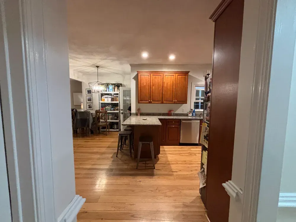 View through a doorway into a kitchen with wood cabinets, island stools, and hardwood floors
