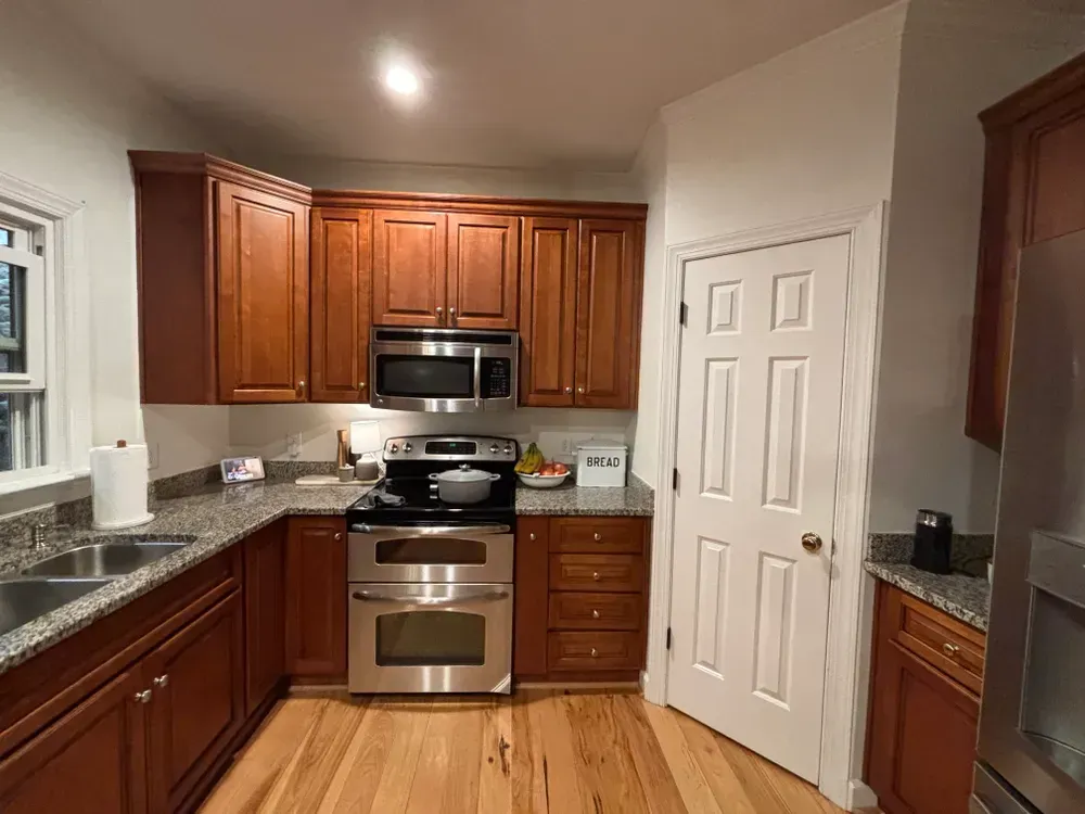 Compact kitchen with wood cabinets, stainless steel appliances, granite counters, and a white pantry door.