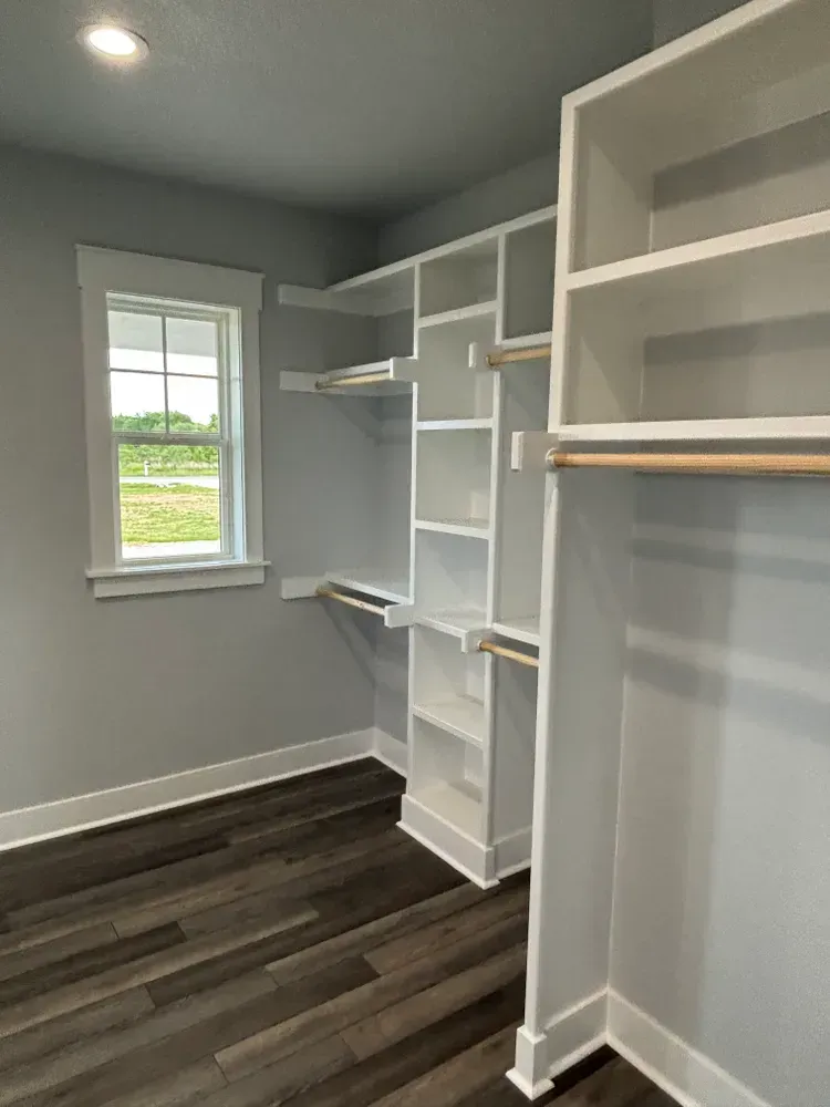 Empty walk-in closet with white shelving, gray walls, dark wood floor, and a window.