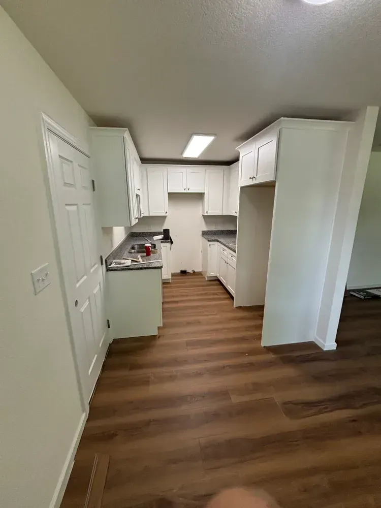 Narrow white kitchen with wood-look flooring, cabinets, and a door on the left.