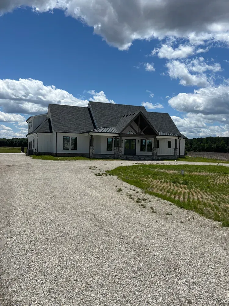 Modern single-story house with dark roof beside a gravel driveway under a cloudy blue sky