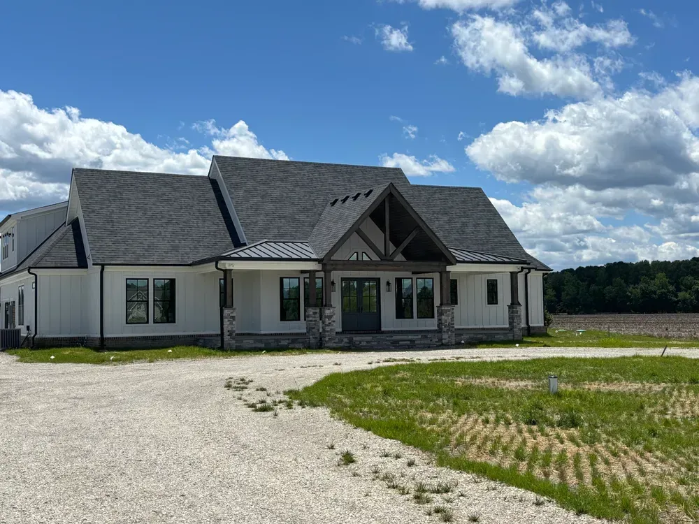 Modern white house with dark gray roof and front porch on a gravel lot under a partly cloudy sky
