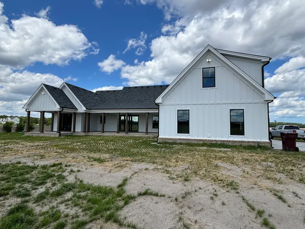 White house under construction on a sandy lot, with a dark roof and cloudy blue sky.