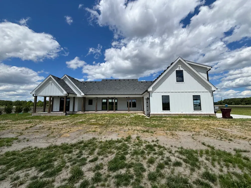 White farmhouse with gray roof and covered porch under a cloudy blue sky, surrounded by a gravel lot and grass.