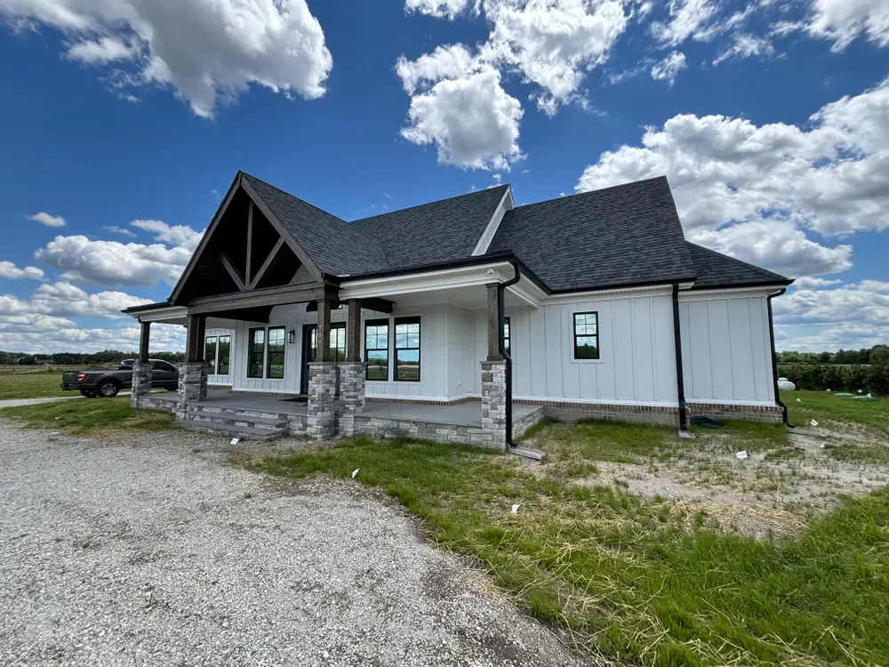 White house with dark roof and porch under a cloudy blue sky, seen from a gravel driveway.