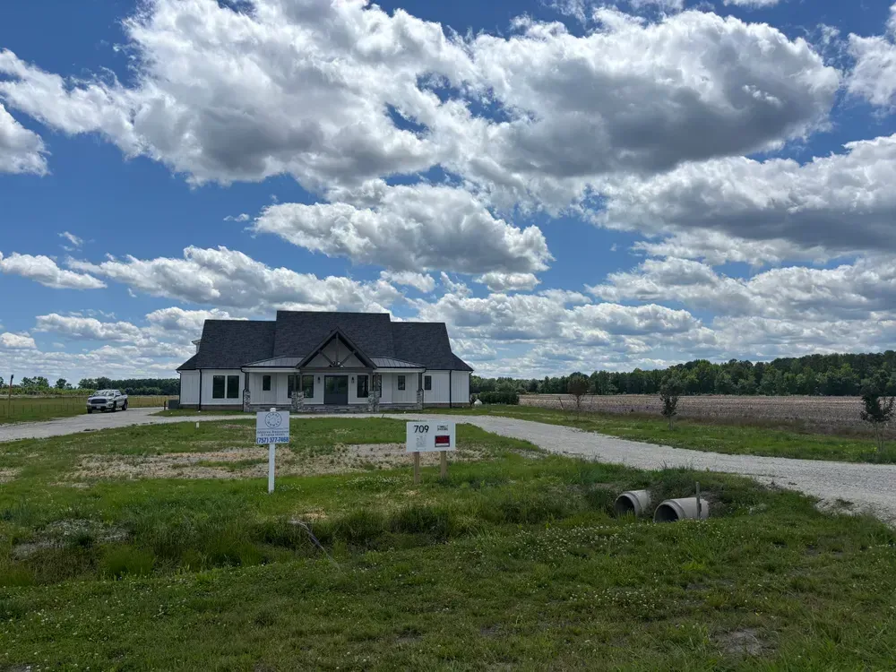 Gray house under a cloudy sky beside a gravel lot and grassy field