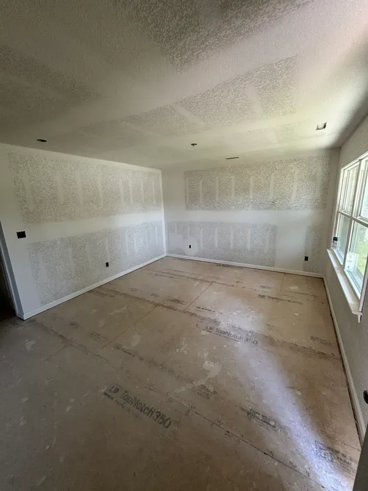 Empty room under renovation with bare drywall, unfinished floor, and large windows on the right.