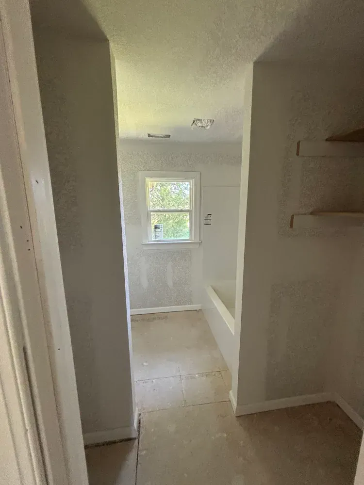 Narrow hallway leading to a bright room with a window, light walls, and beige tile floor.