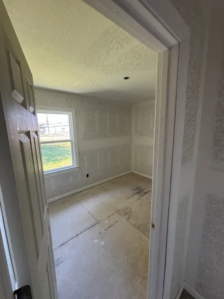 Small empty room with white walls, a window, and unfinished flooring, viewed through a doorway