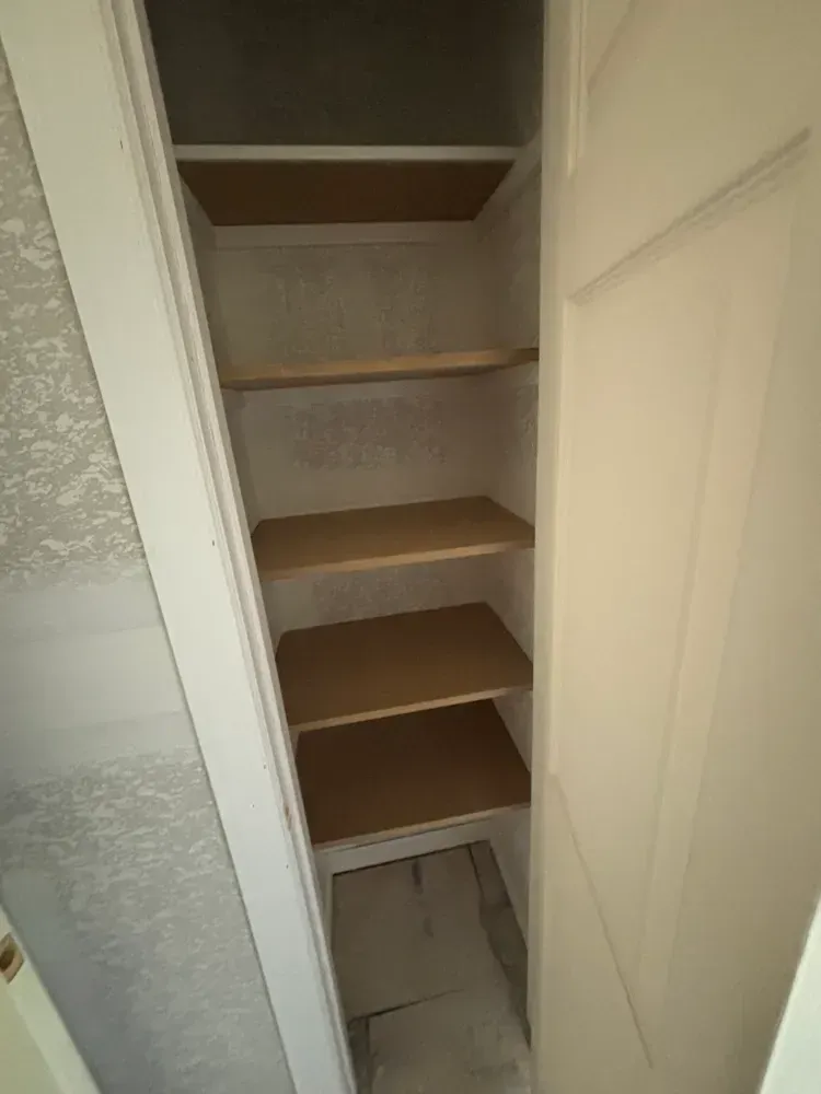 Empty narrow pantry closet with built-in wooden shelves and open white doors.