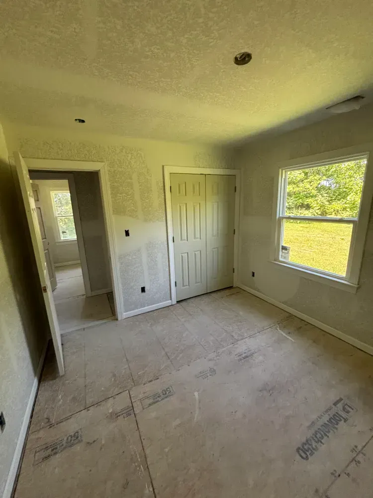 Empty bedroom under renovation with bare drywall, white closet doors, and a window letting in sunlight.