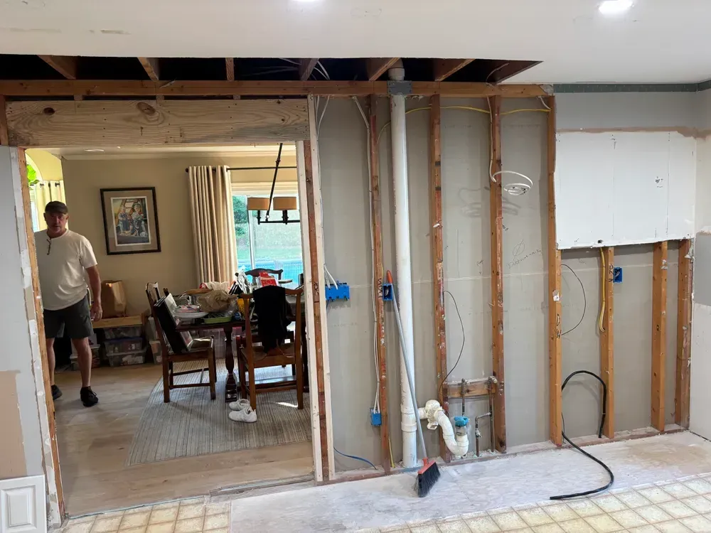 Partially demolished interior wall revealing studs, wiring, and a dining room beyond