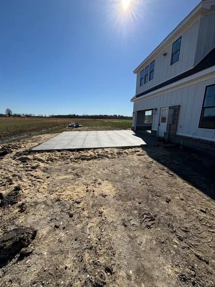 Sunlit farmhouse exterior with dirt yard and concrete slab beside open fields
