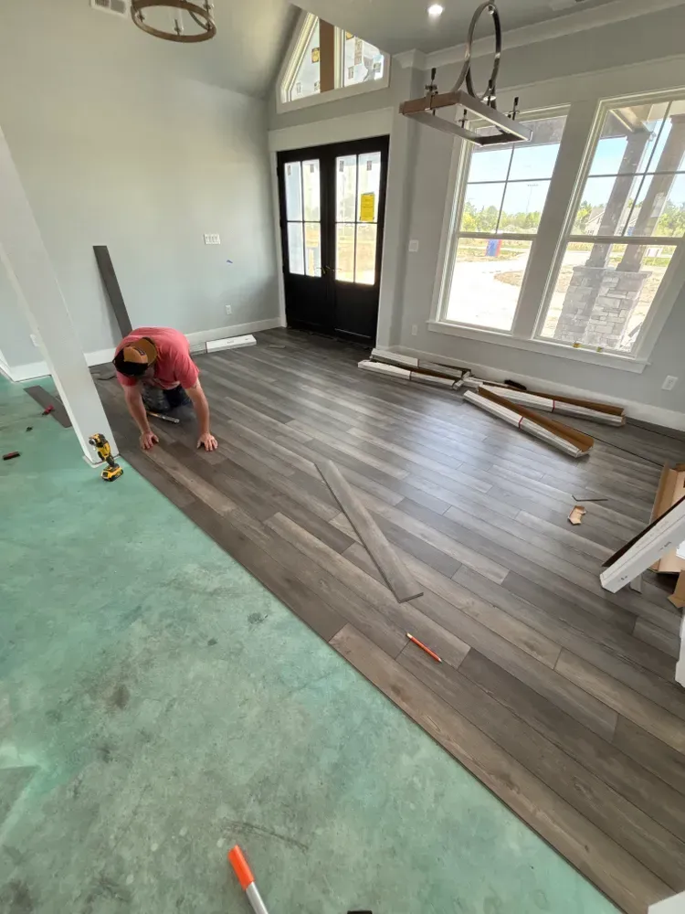 Person installing gray wood flooring in a bright room with large windows and unfinished floor areas