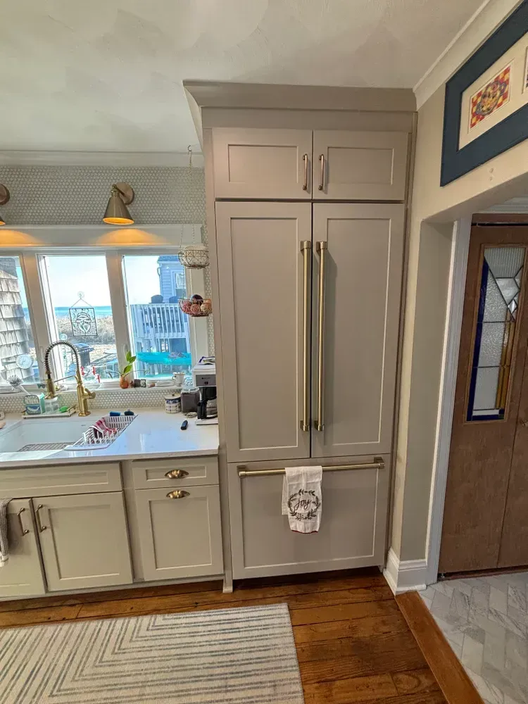 Bright kitchen with tall beige refrigerator cabinets, sink under window, and wooden floor.