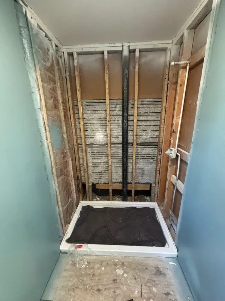 A narrow bathroom under renovation with exposed studs, unfinished walls, and a shower base covered in debris.