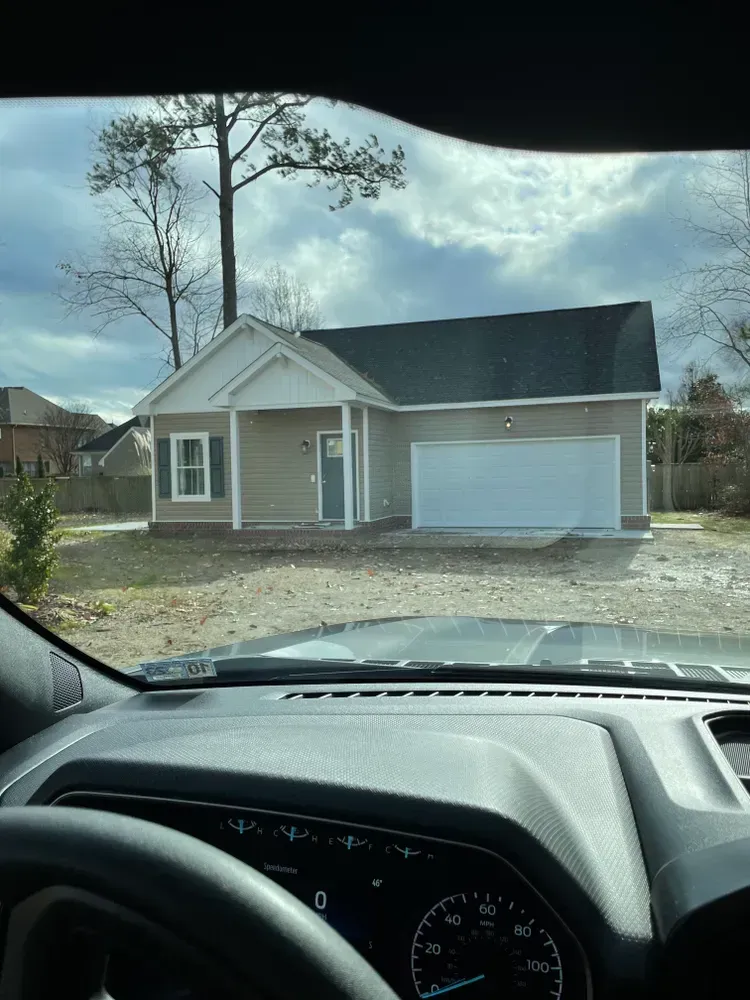 View from a car of a small beige house with a white garage and bare trees in front