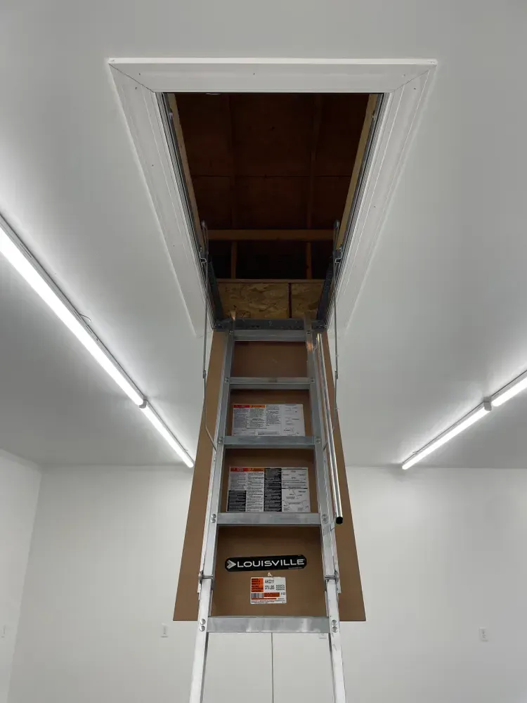 Ladder extended to an open attic hatch in a white room with fluorescent lights.