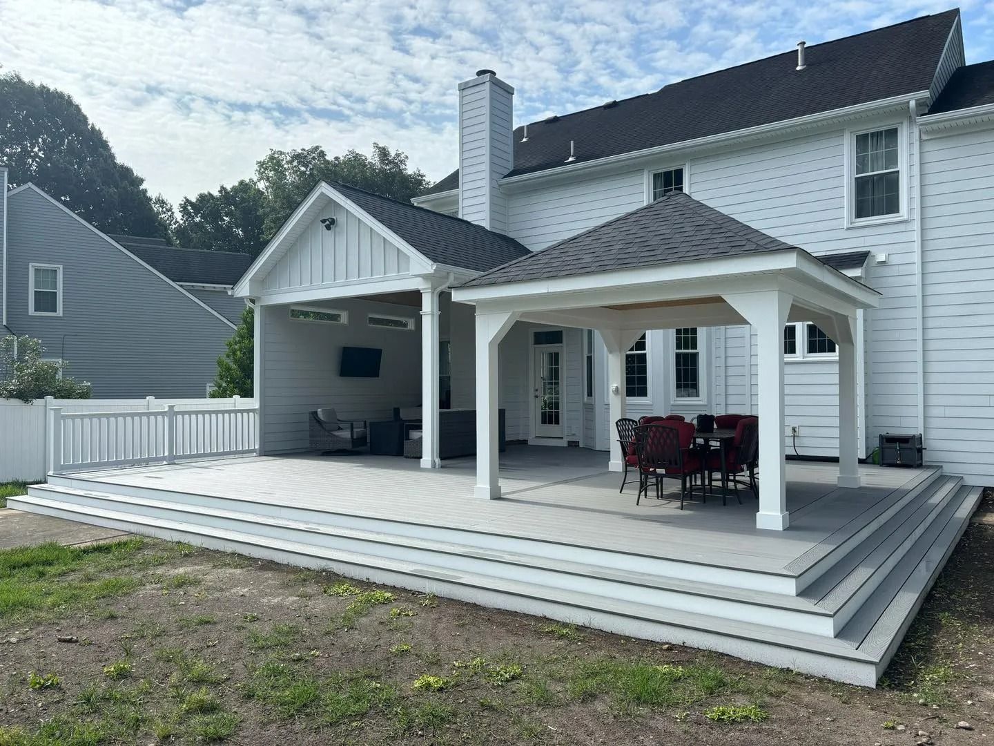 White house with a large wooden deck, covered patio, and outdoor dining area in the backyard.