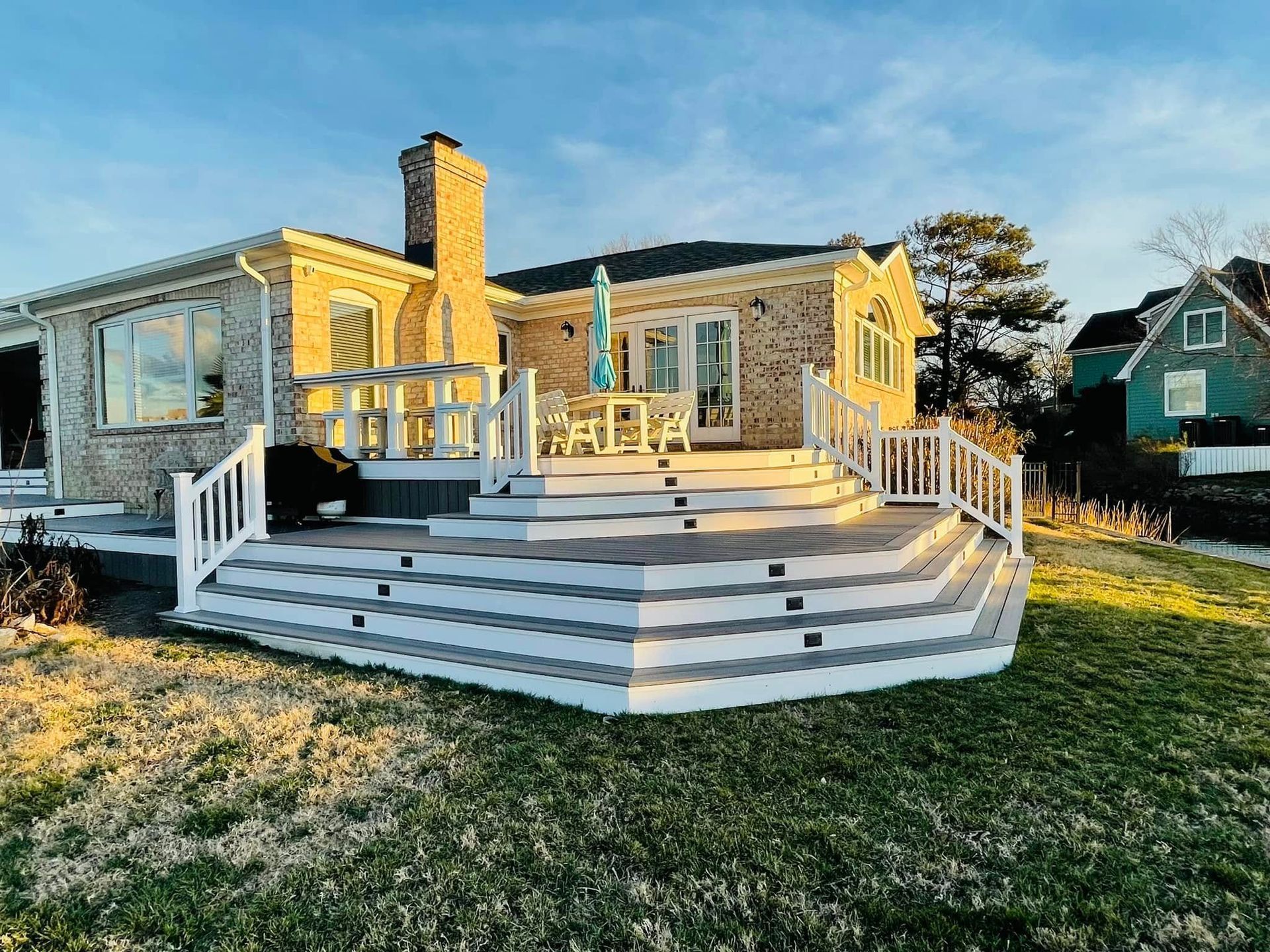 Single-story house with a white wooden deck and railings in a sunny yard at dusk.