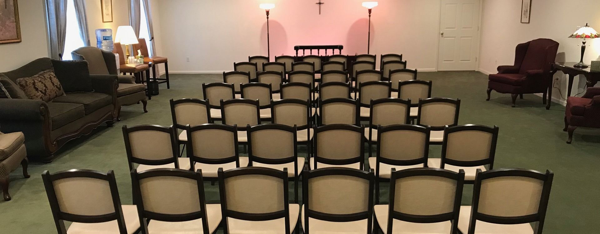 Rows of chairs face a small table and crucifix in a room with couches and lamps.