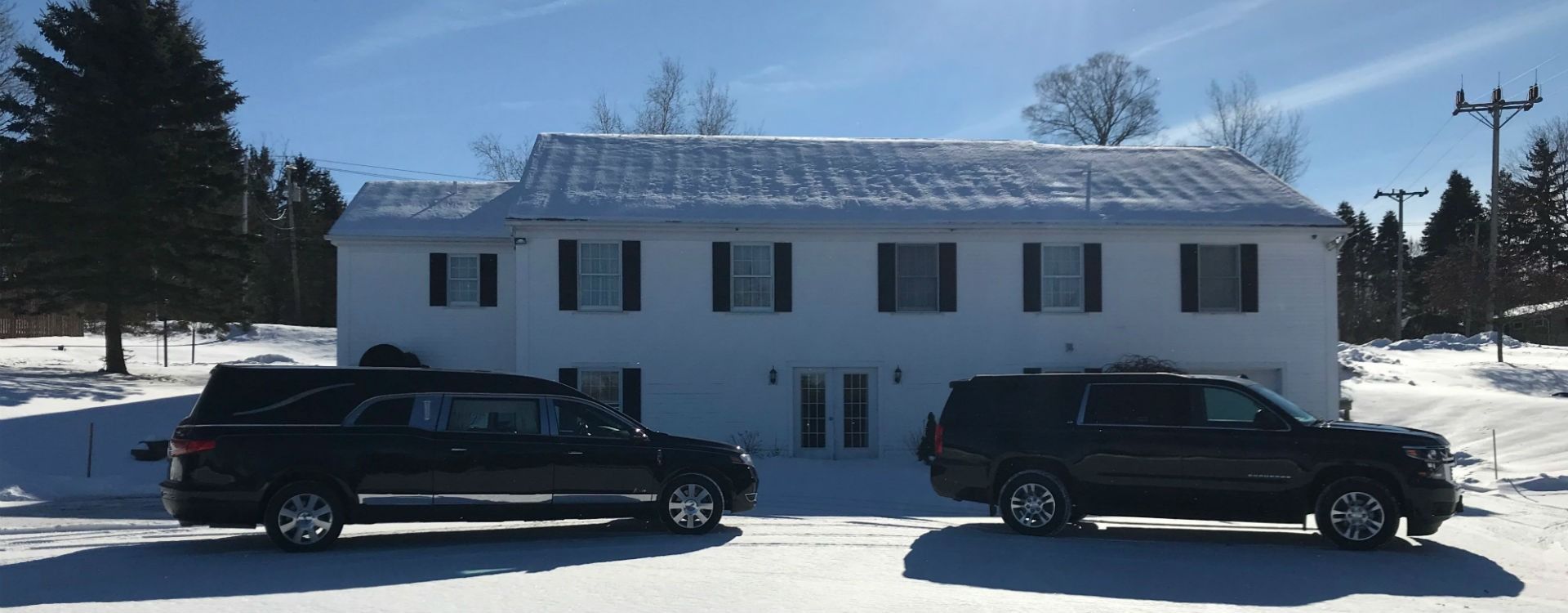 Hearse and SUV parked in front of a white building with snow on the roof. Snowy ground and trees.