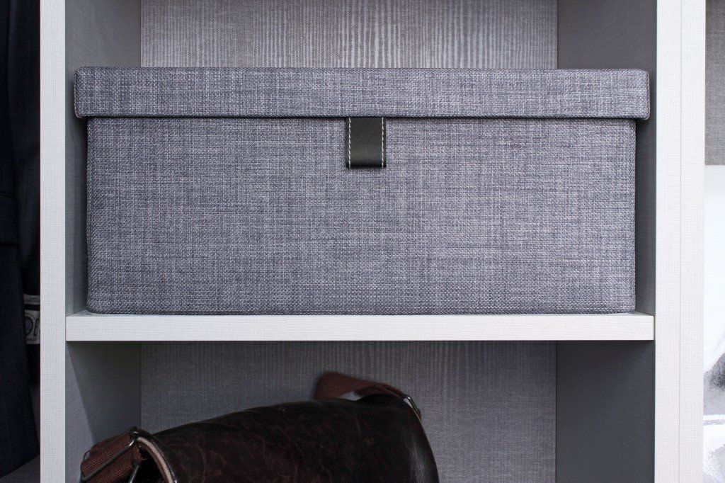 Gray storage box with a metal clasp on a shelf in a closet. A brown purse is below.