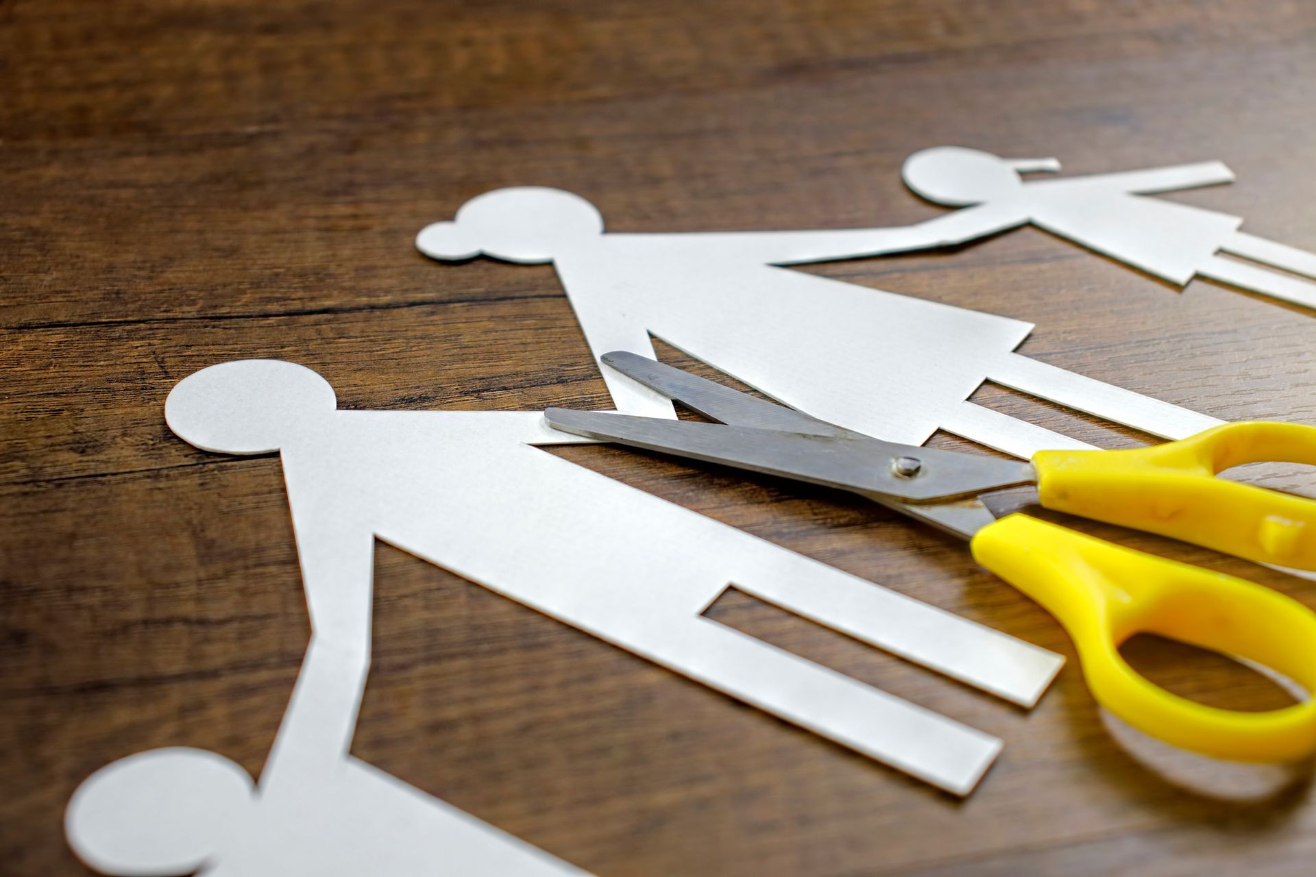 A pair of scissors is cutting a paper family on a wooden table.