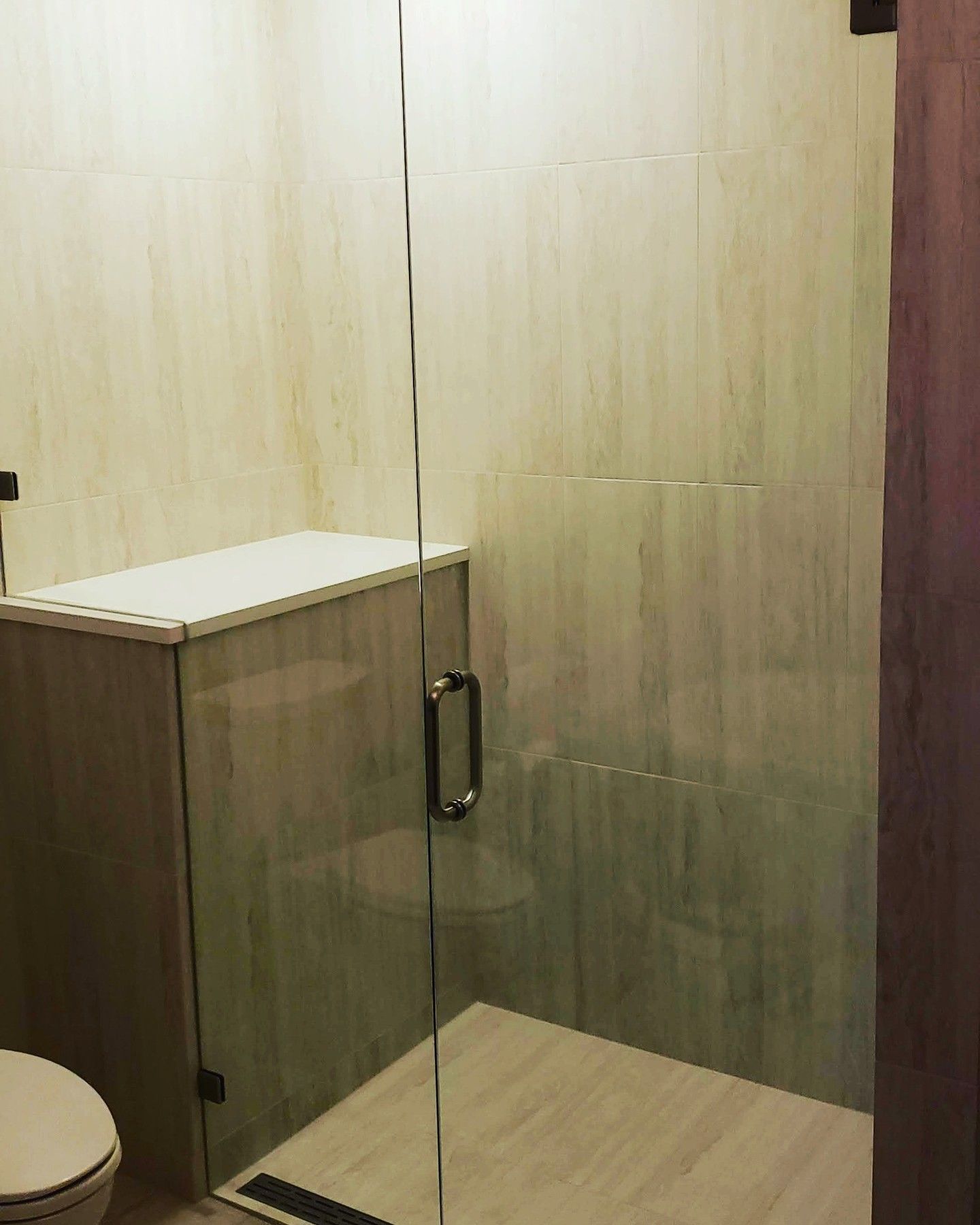 Glass-enclosed shower with beige tile walls and flooring, a dark shower drain, and a dark cabinet with a white top.