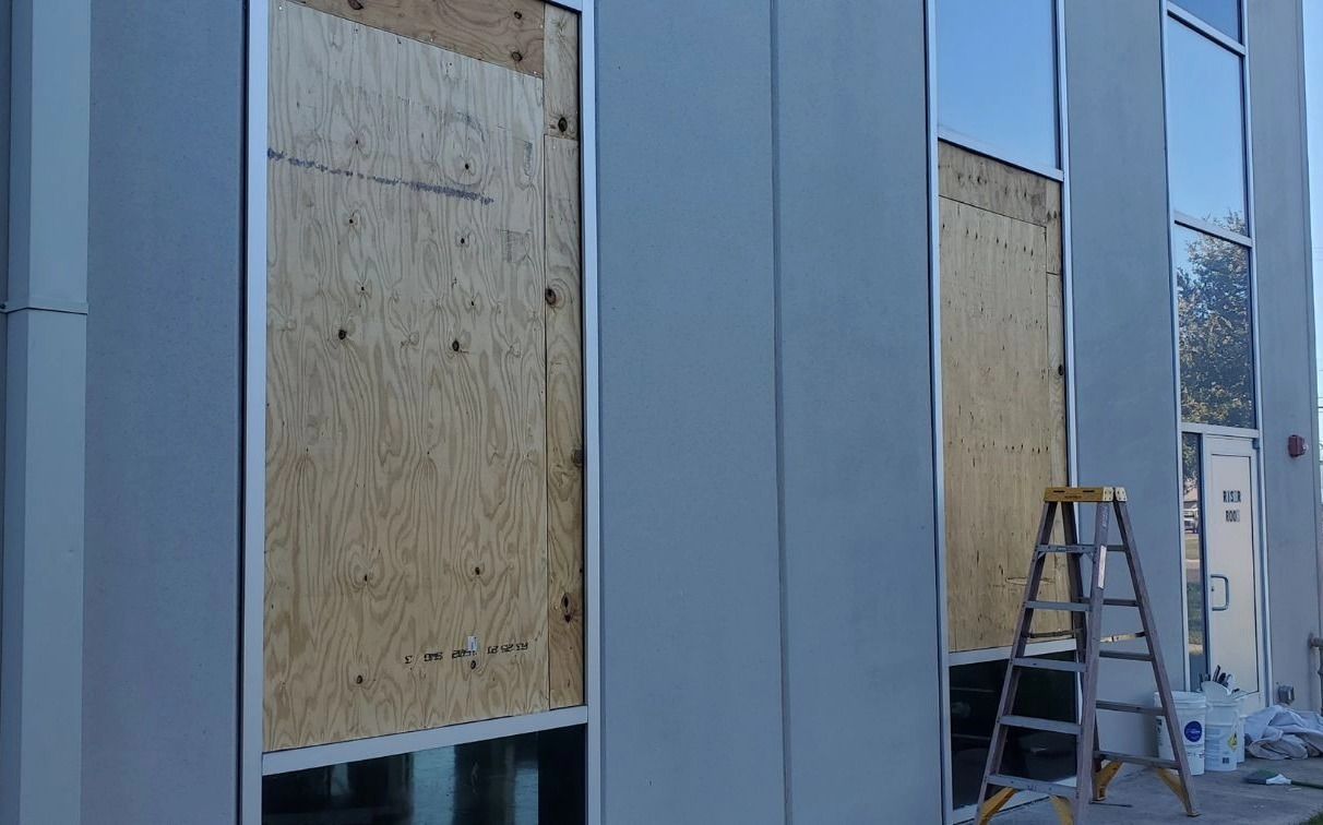 Exterior of a building with plywood covering some windows, a ladder propped against a window, and light blue siding.