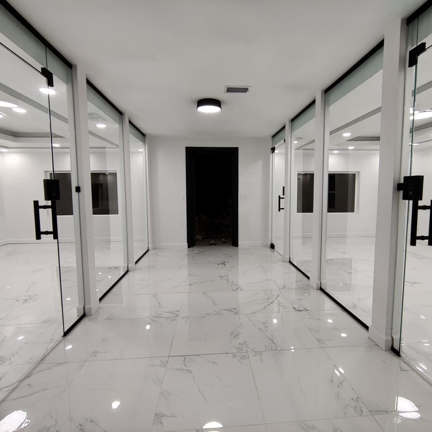 A modern office hallway with glass-walled rooms. White marble floors lead to a dark doorway at the end.