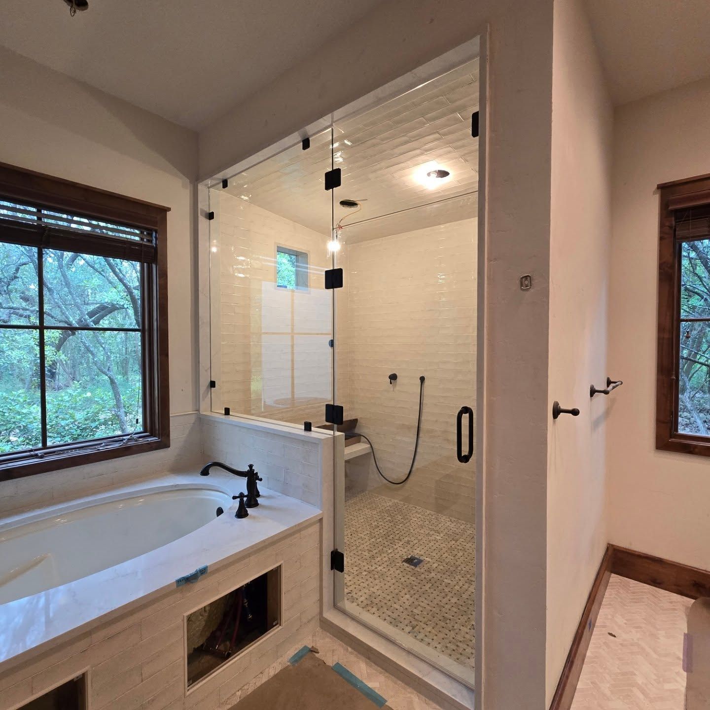 A modern bathroom featuring a glass shower with black fixtures, a bathtub, and two windows with wooded views.