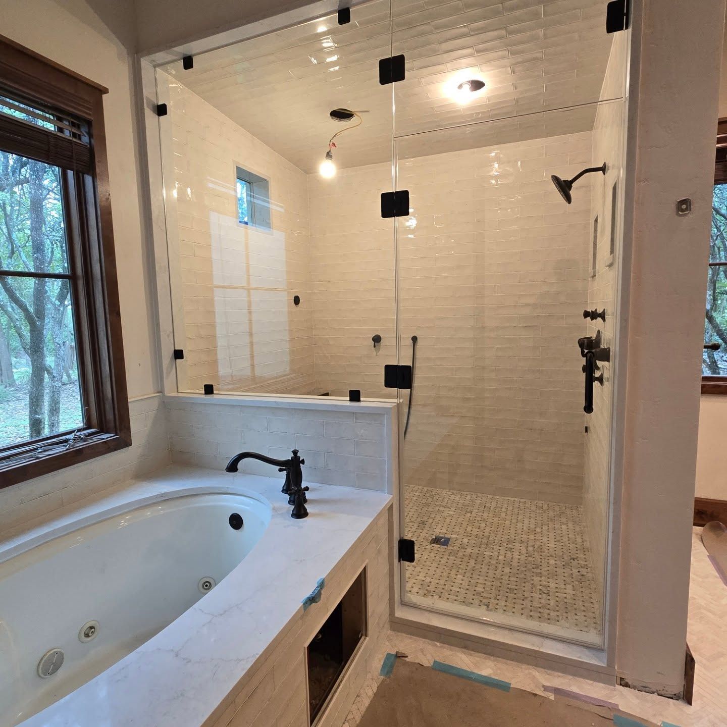 Bathroom with a white bathtub, glass shower enclosure, and beige tiled walls. Black fixtures and window with a forest view.