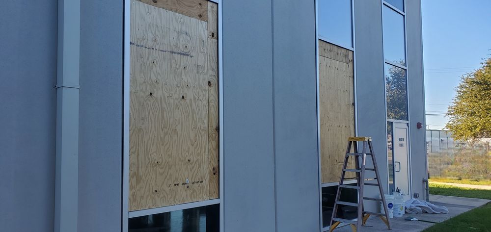 Exterior of a building with large windows, some boarded up with plywood. A ladder leans against the building.