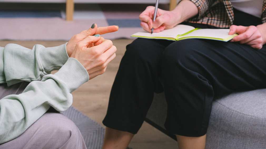 A woman is sitting on a couch talking to a man who is writing in a notebook.
