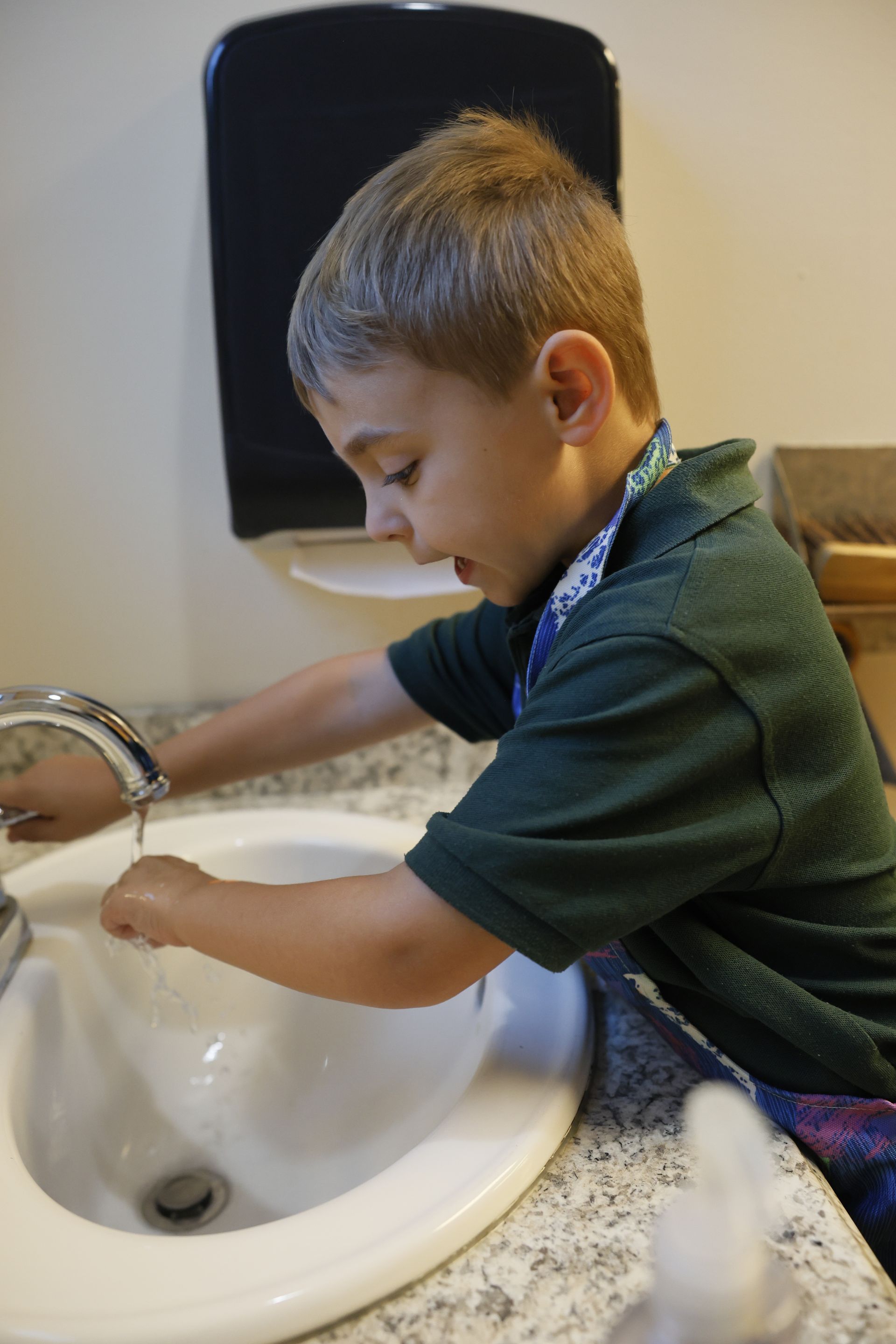 Child washing hands at a white sink in a bathroom, with soap dispenser above.