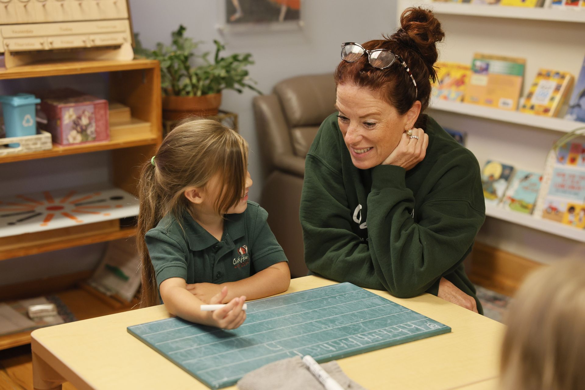 A Montessori guide smiles at a child in a Montessori classroom. The child looks up with a white chalk.