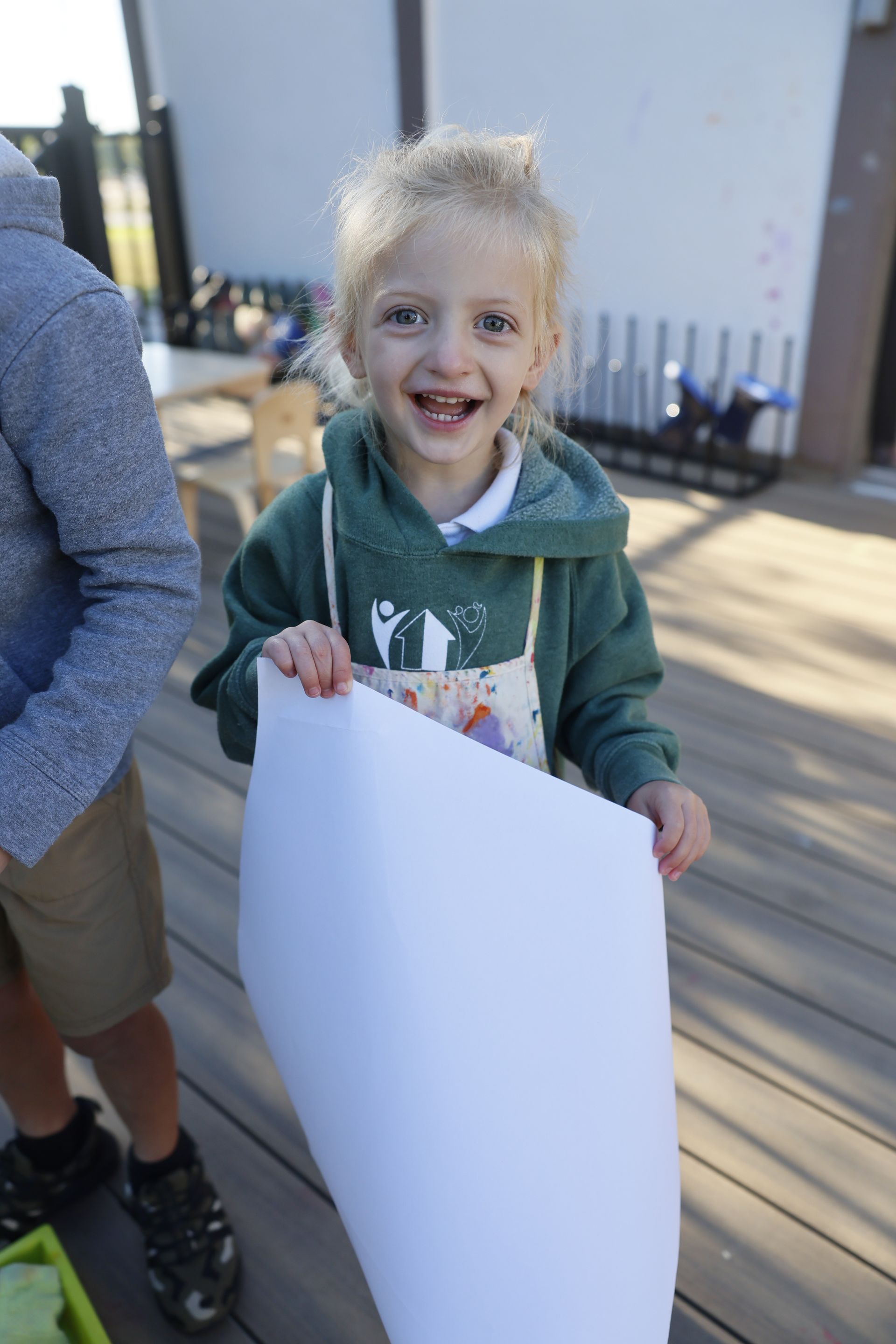 Smiling Montessori child in a green hoodie holding a large white paper outdoors on a sunny day.