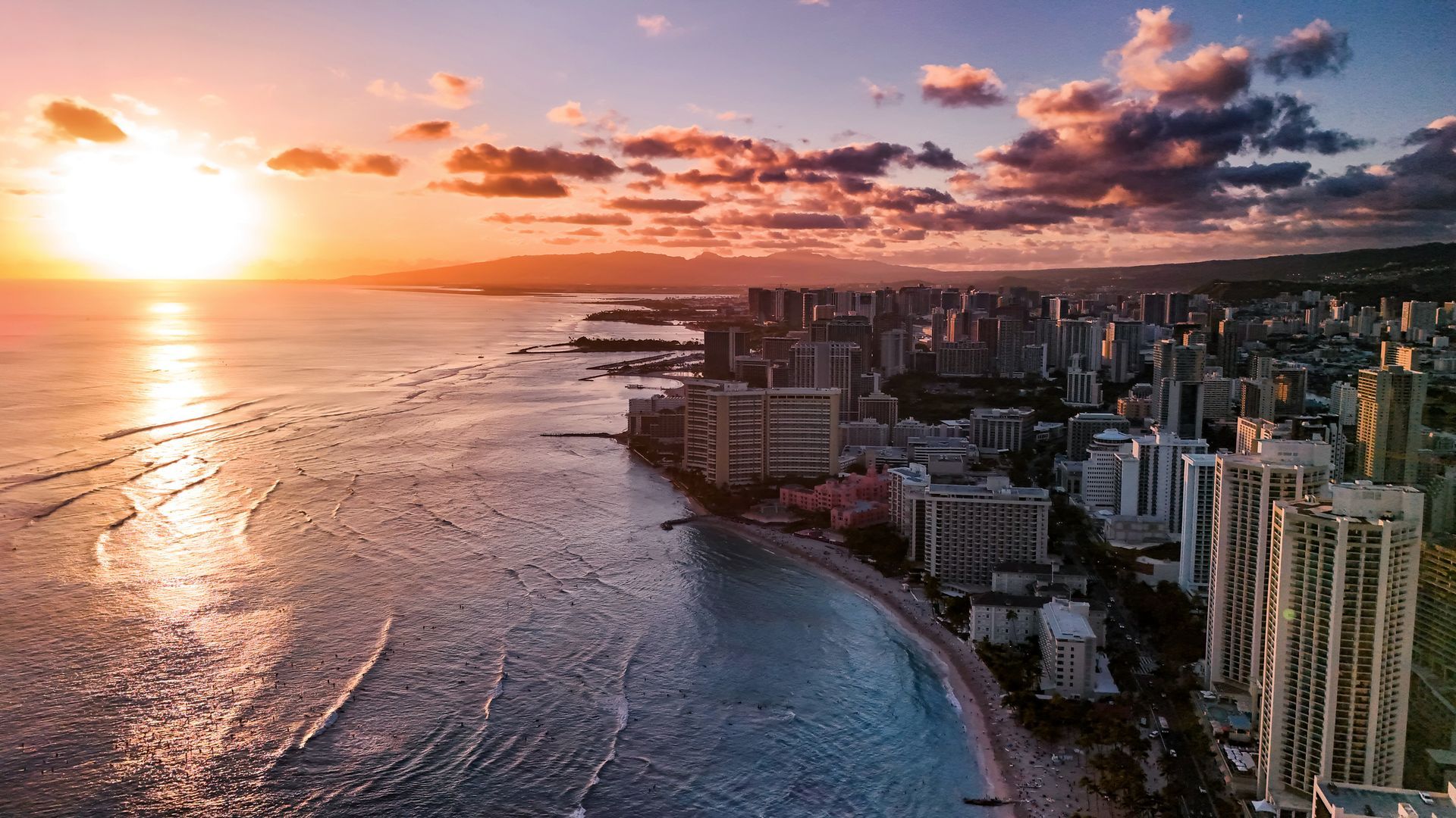 Sunset over Waikiki Beach, Hawaii, with cityscape and ocean waves, vibrant orange and purple colors.
