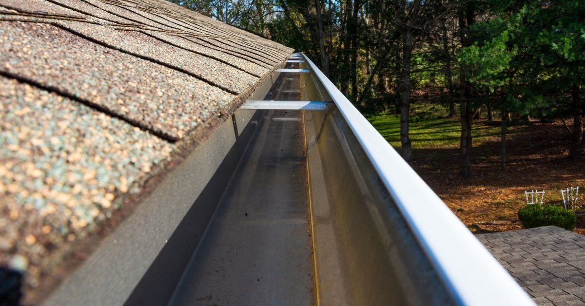 A close-up of a clean gutter on a house, with a brick patio and lawn with many trees beyond it.