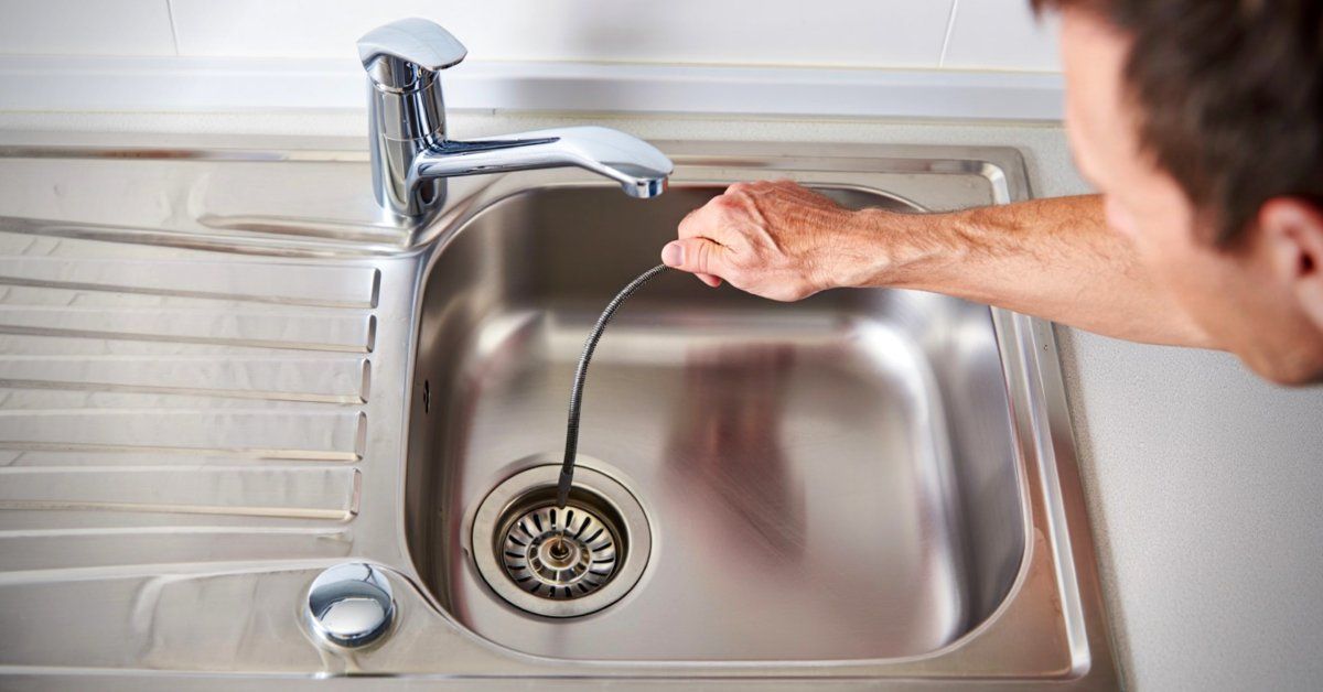 A man getting ready to use a drain snake in his kitchen sink. He's holding the snake above the drain