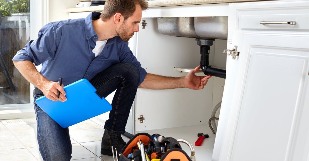 A plumber crouched down, looking at the underside of a sink. He's taken off the cabinet door and is reaching for a black pipe.