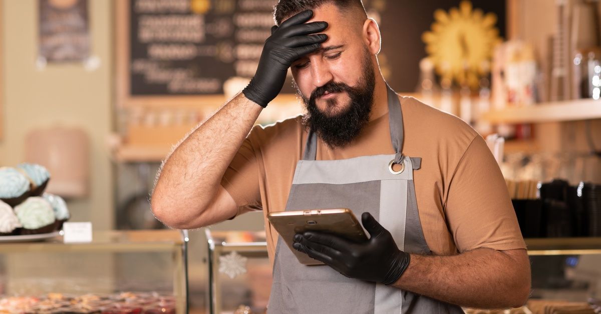 A restaurant owner looking at a tablet in his hand. His other hand is on his forehead. There are many pastries behind him.