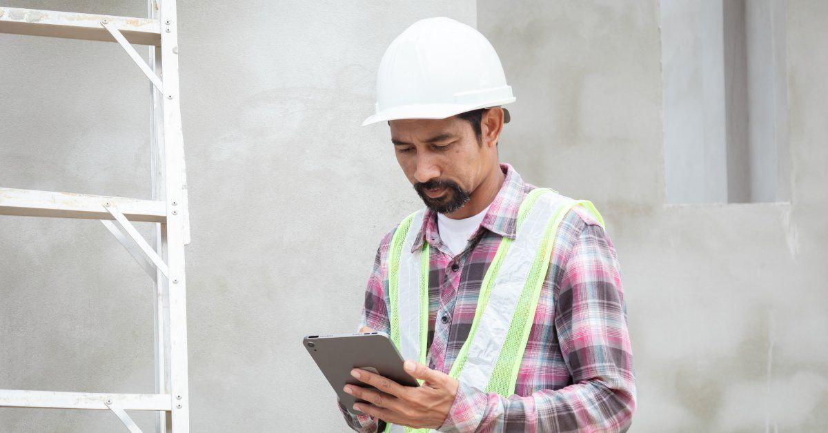 A man in a hard hat and vest looking at the tablet in his hands. There is a ladder propped up against the wall next to him.
