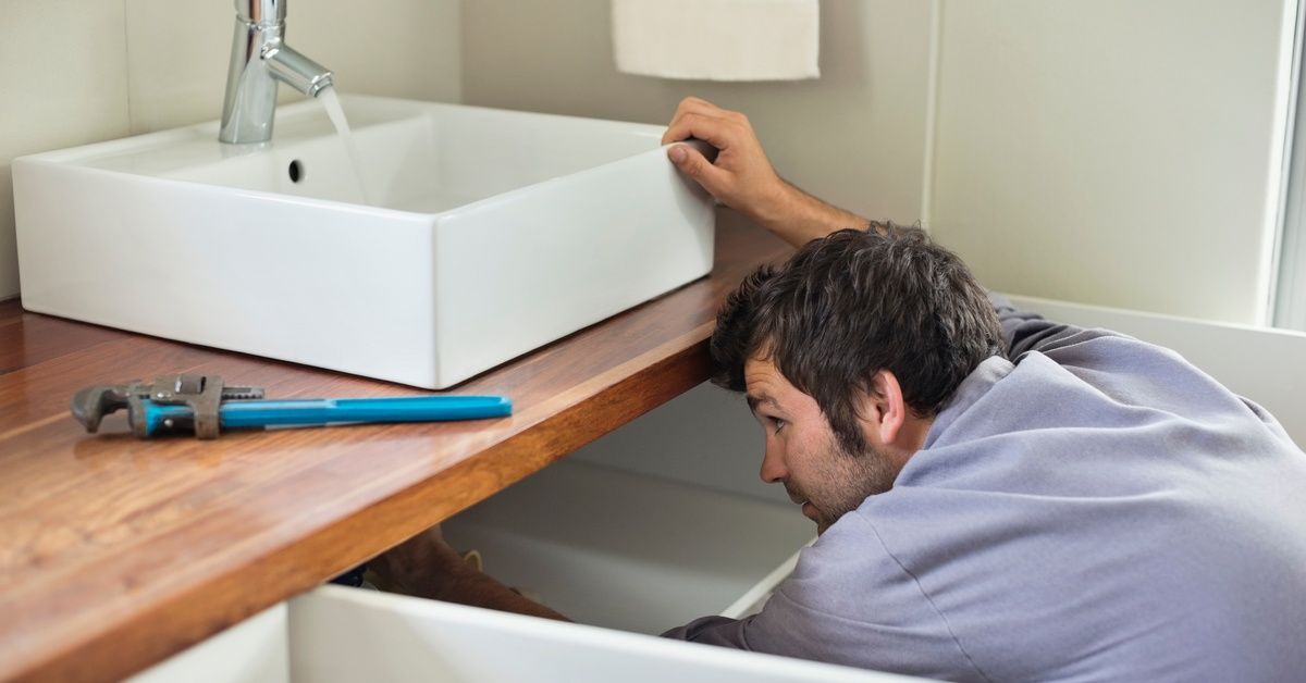 A man looking underneath his bathroom sink while the water is running. There is a blue wrench sittin