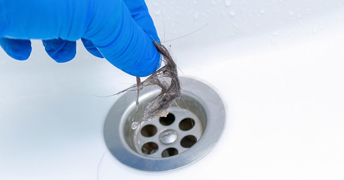 A gloved hand pulling a clump of wet hair out of a drain. The the person is wearing a blue glove.